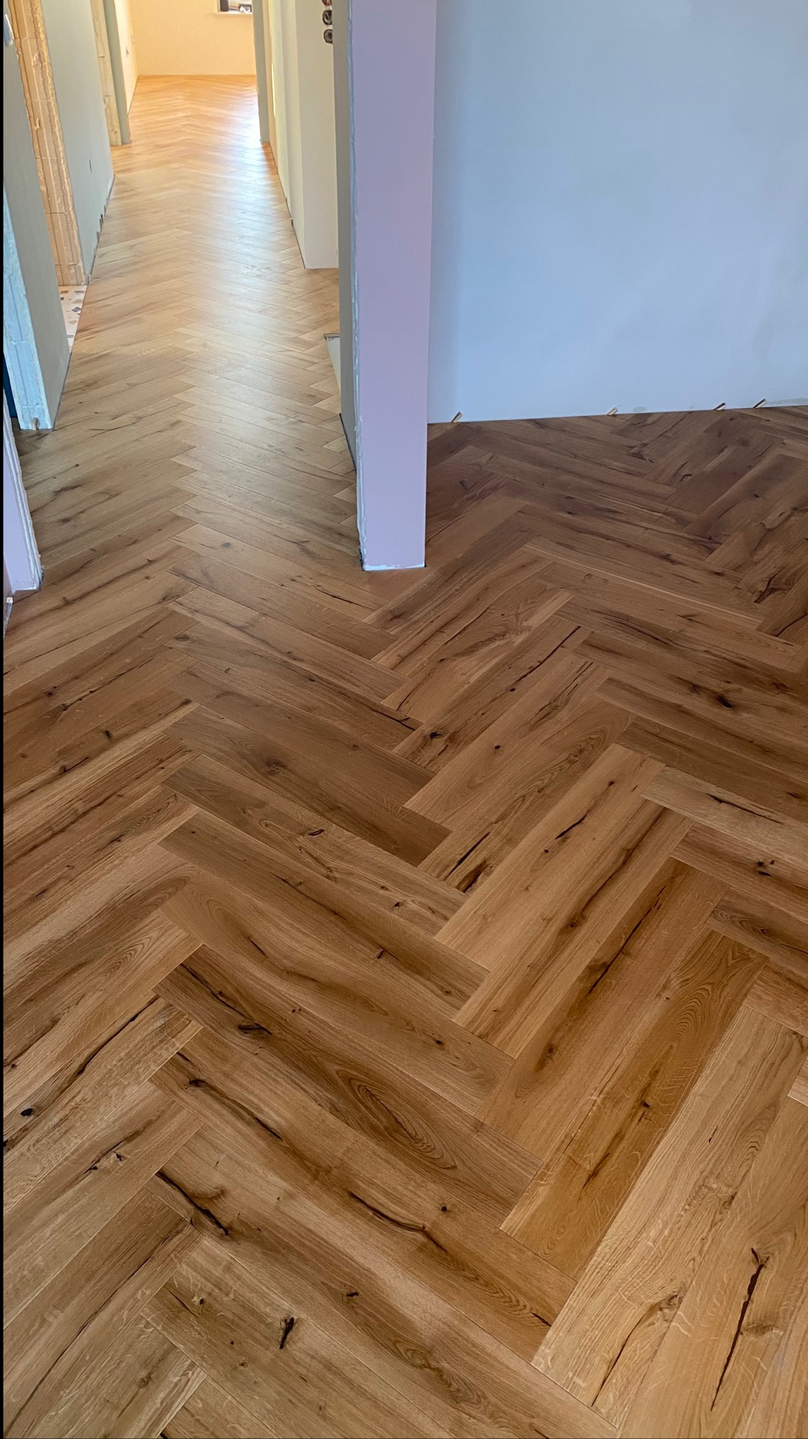 Ukończony parkiet ułożony we wzór jodełki francuskiej, widoczne naturalne usłojenie drewna, w tle fragment korytarza z białymi ścianami i pomalowanym na jasny fiolet filarem.