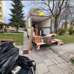 Otwarta paka busa załadowana kartonami i meblami podczas przeprowadzki. Na chodniku czarne worki i paczka słodyczy. Tło: budynki mieszkalne, drzewa.