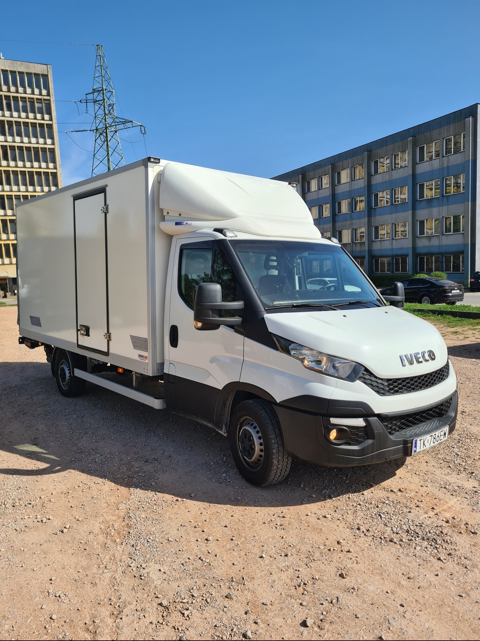 Biały furgon Iveco z zabudową kontenerową na żwirowym placu, budynki biurowe w tle, błękitne niebo. Transport towarów, przeprowadzki.