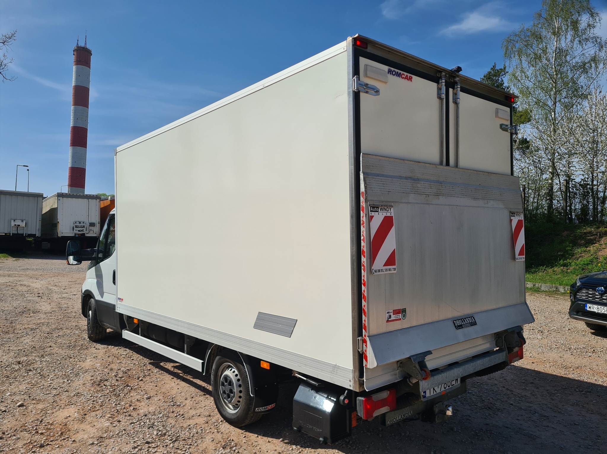 Biały samochód dostawczy z otwartą windą załadowczą marki DHOLLANDIA, zaparkowany na żwirowym placu, z kominem w tle.