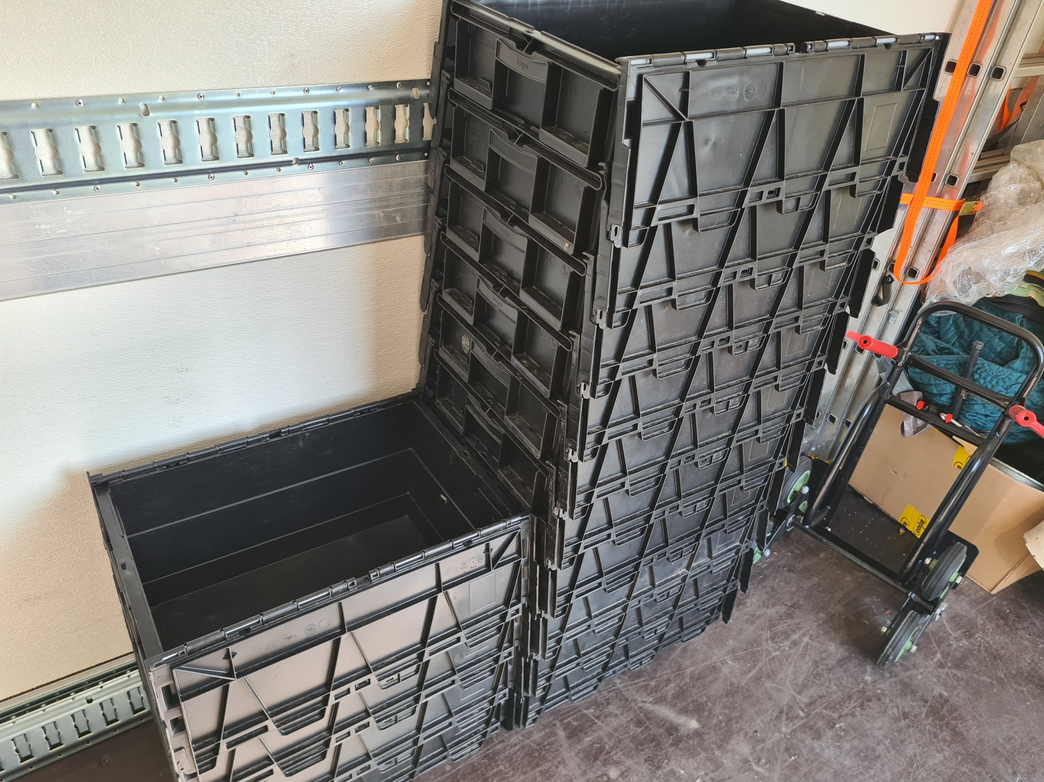 Stos czarnych, plastikowych pojemników do przeprowadzki, ustawionych w magazynie, z metalową listwą mocującą widoczną na ścianie i wózkiem transportowym obok.