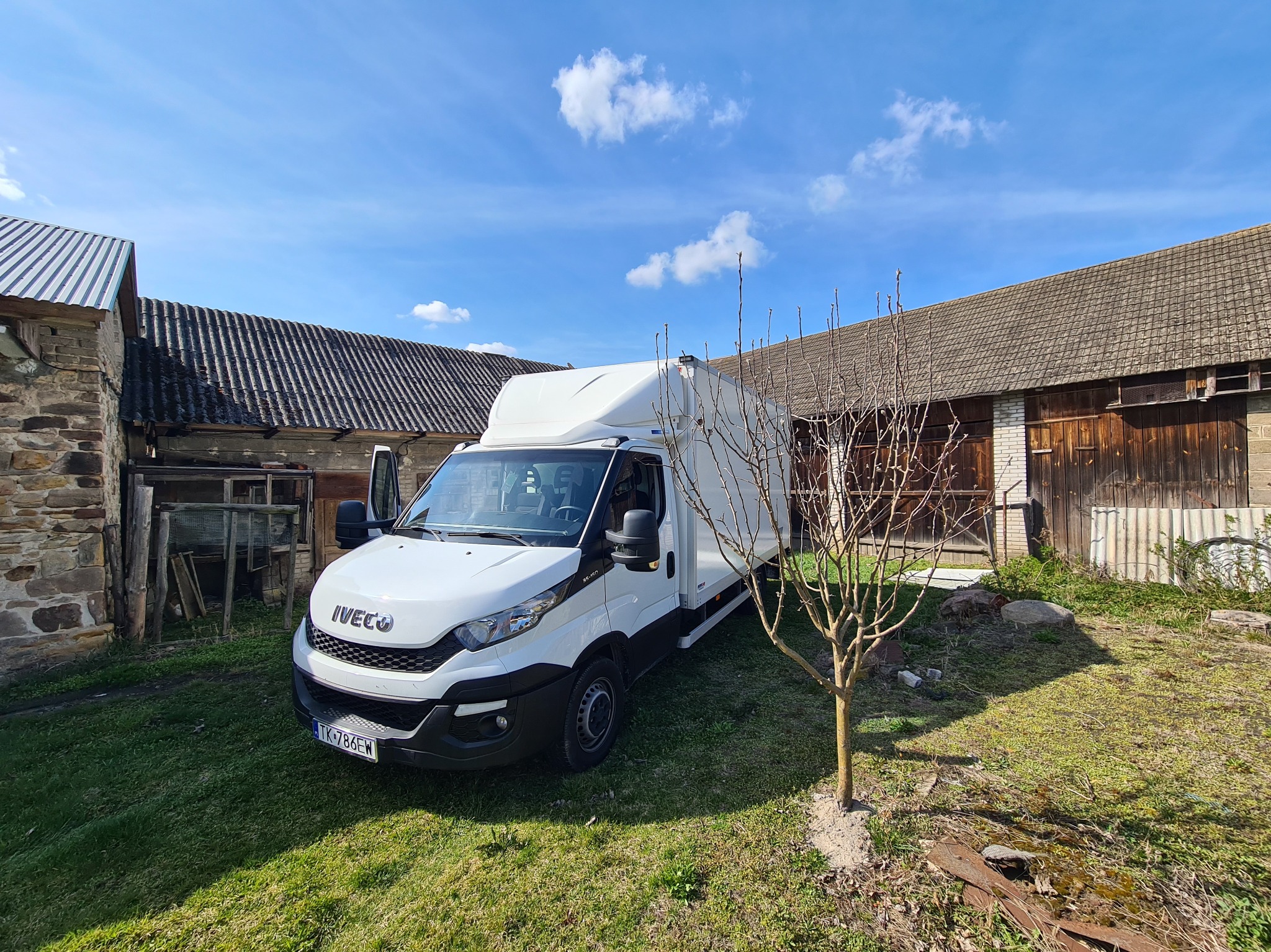 Biały furgon Iveco na trawniku przed starym, drewnianym budynkiem gospodarczym w słoneczny dzień. Otwarta kabina kierowcy.