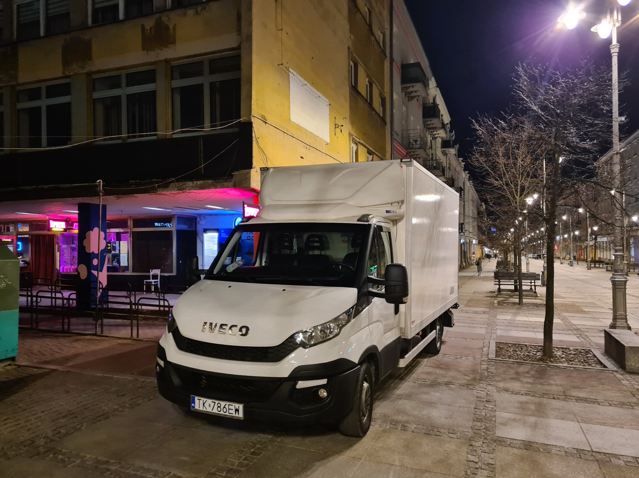 Biały samochód dostawczy Iveco na tle nocnego miasta z oświetlonymi budynkami i latarniami. Widoczny fragment chodnika z kostki brukowej i drzewo.