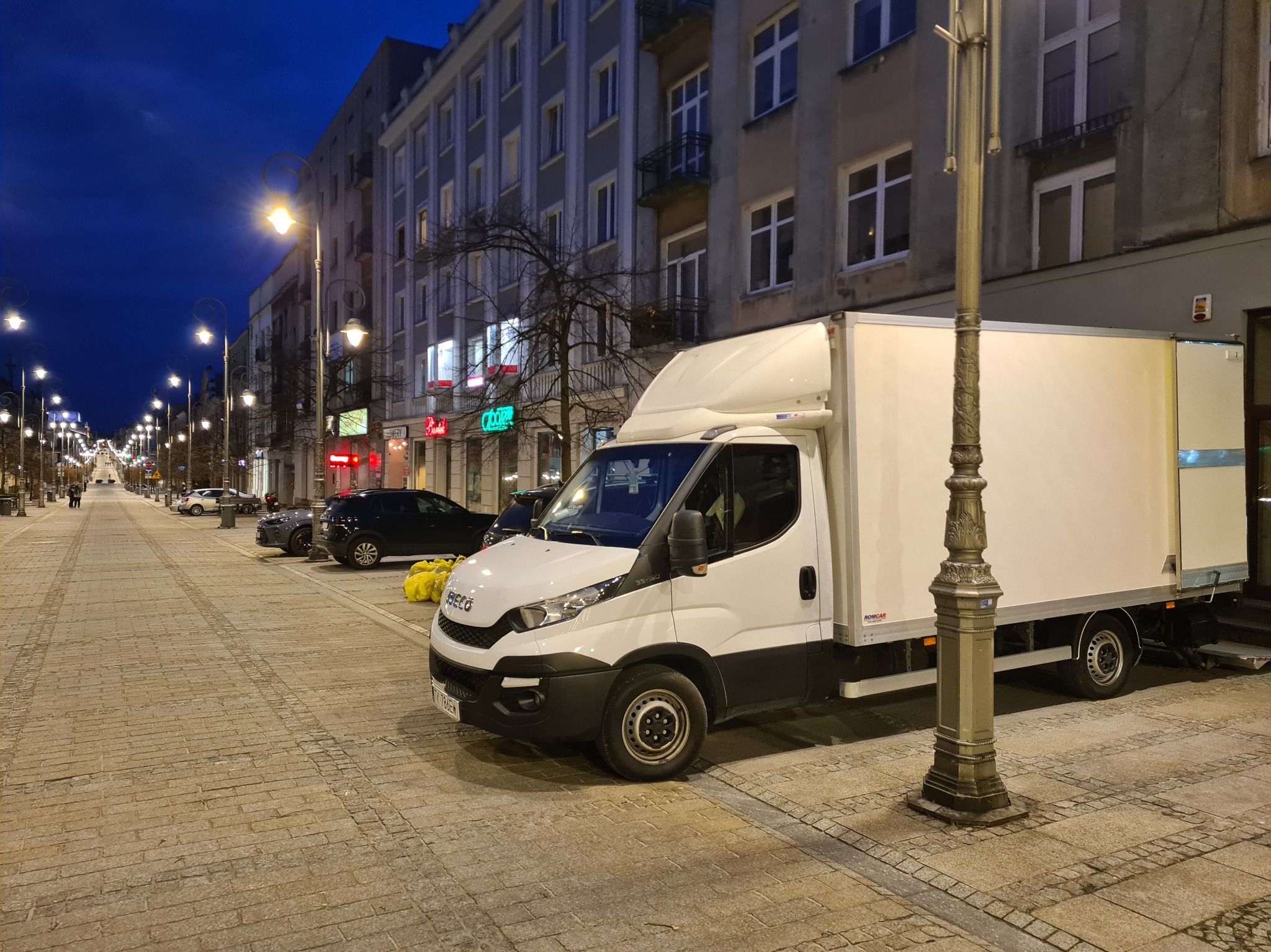 Biały samochód dostawczy Iveco zaparkowany na brukowanej ulicy miasta nocą, oświetlony latarniami. W tle budynki i inne zaparkowane pojazdy.