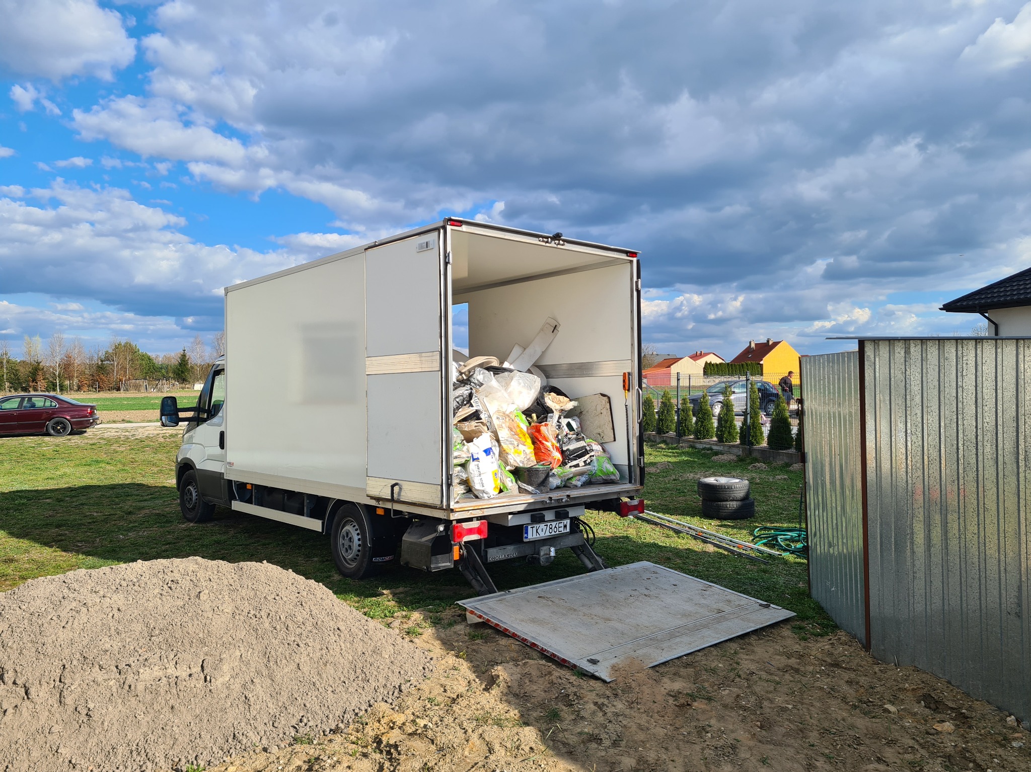 Otwarty biały furgon z platformą załadowaną workami i przedmiotami, zaparkowany na zewnątrz, obok sterty piasku i blaszanej ściany. Błękitne niebo z chmurami w tle.