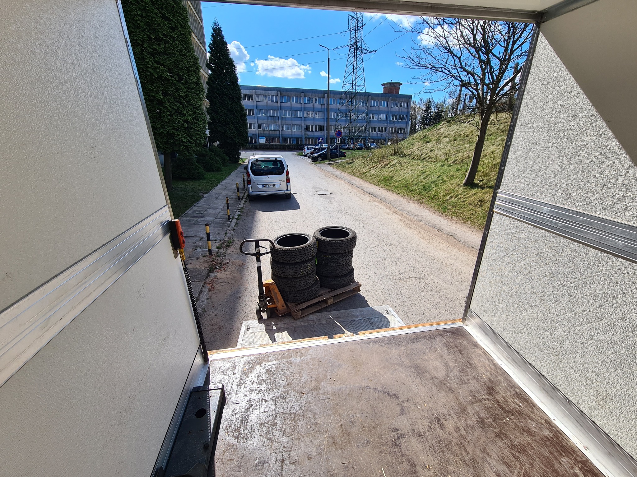 Wnętrze busa z paletą opon na wózku paletowym, widok na ulicę z budynkiem w tle, jasny dzień.