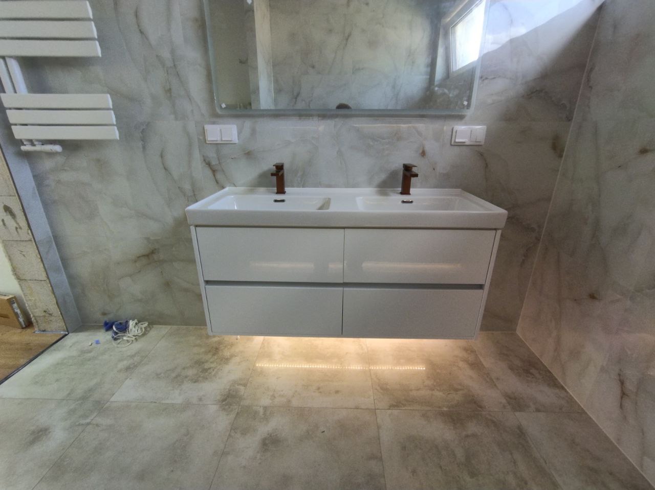 Nowoczesna łazienka z podwójną białą umywalką nablatową, baterie w kolorze miedzi, podświetlenie LED pod szafką i marmurowe płytki na ścianach.