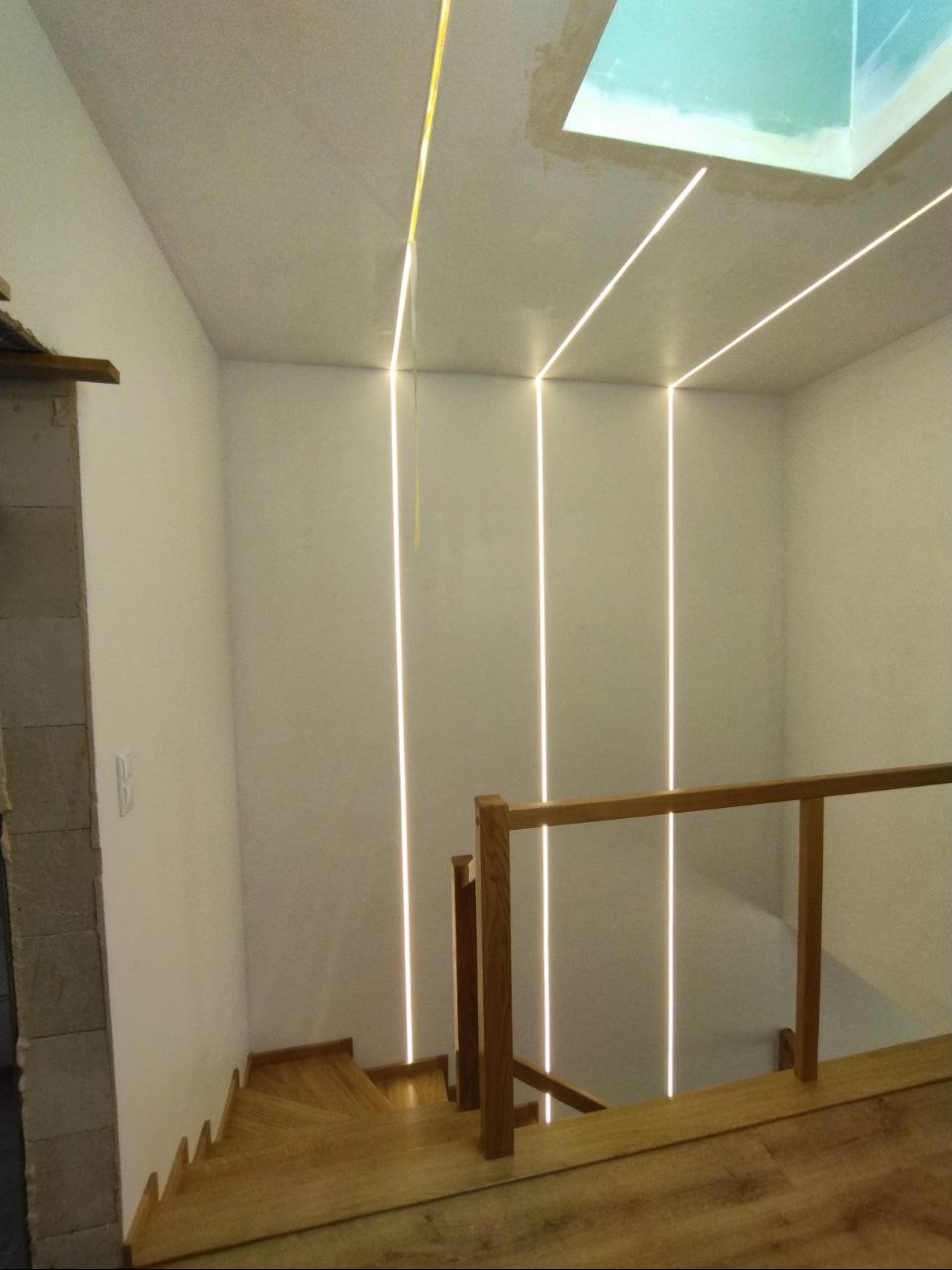 Wnętrze klatki schodowej z drewnianymi schodami i balustradą, oświetlone pionowymi liniami LED wpuszczonymi w ściany i sufit, z widocznym fragmentem okna dachowego.