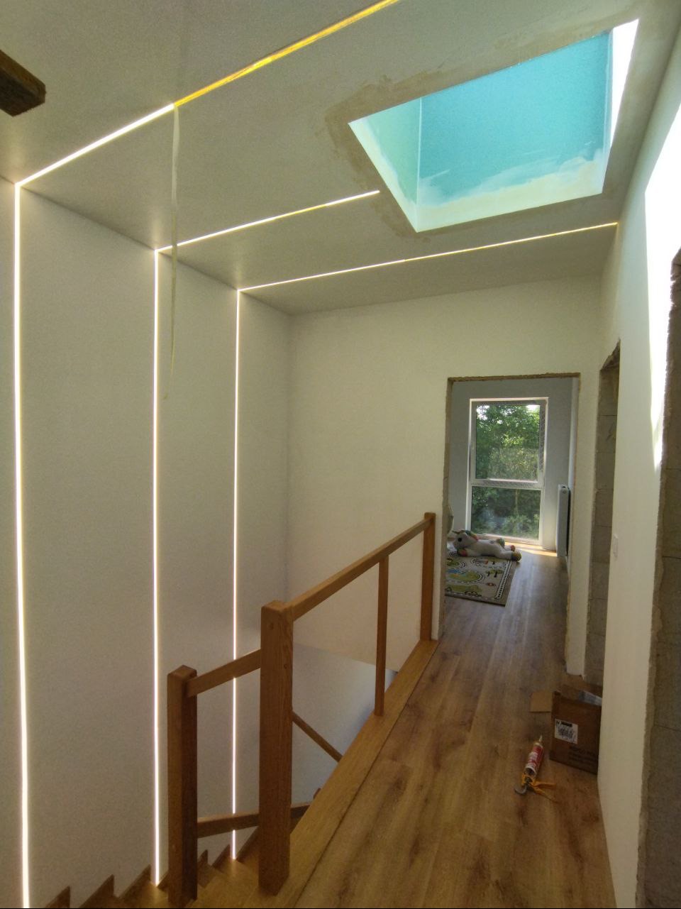 Korytarz z drewnianymi schodami i balustradą oświetlony liniowymi lampami LED w ścianach i suficie, dodatkowo doświetlony oknem dachowym z widokiem na niebo.