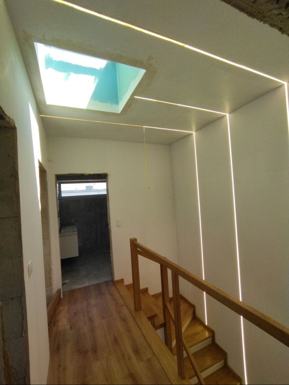 Klatka schodowa z drewnianymi schodami i poręczą, oświetlona liniowymi lampami LED w suficie i ścianach, widok na okno dachowe i wnętrze łazienki w tle.