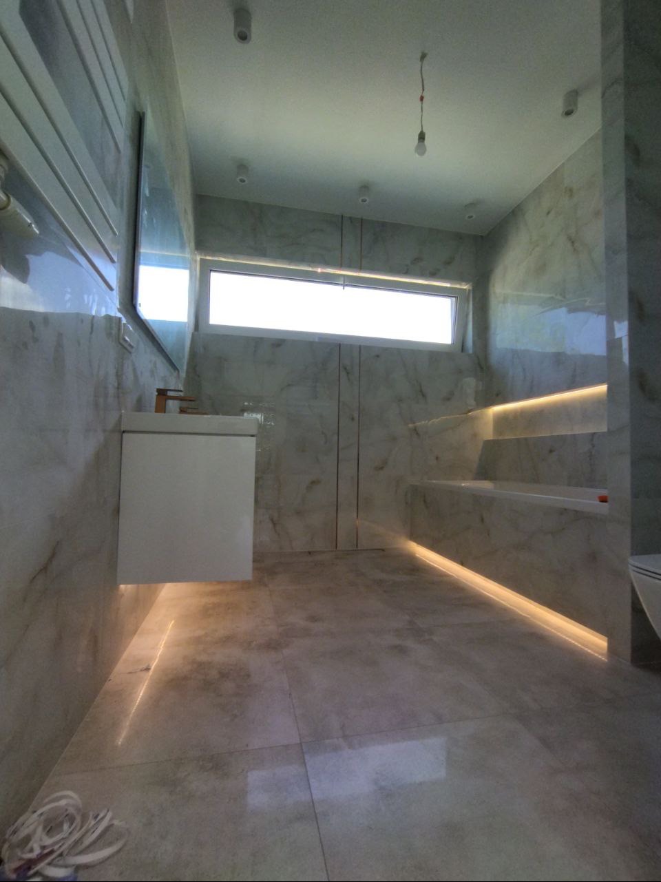 Nowoczesna łazienka z marmurowymi płytkami, oświetleniem LED wzdłuż podłogi i wnęk, wiszącą szafką z umywalką i minimalistyczną armaturą, oraz żarówką wiszącą na kablu zamiast lampy.