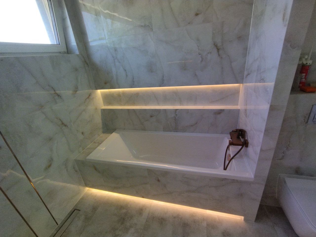 Nowoczesna łazienka z wanną obudowaną marmurem, podświetleniem LED pod półkami i wzdłuż podłogi, oraz designerską, miedzianą baterią.