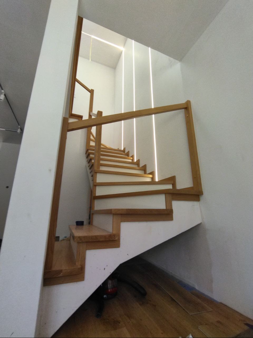 Drewniane schody z białymi podstopnicami i modernistycznym oświetleniem pionowym na ścianie, widok z dołu, w tle widoczne narzędzia.