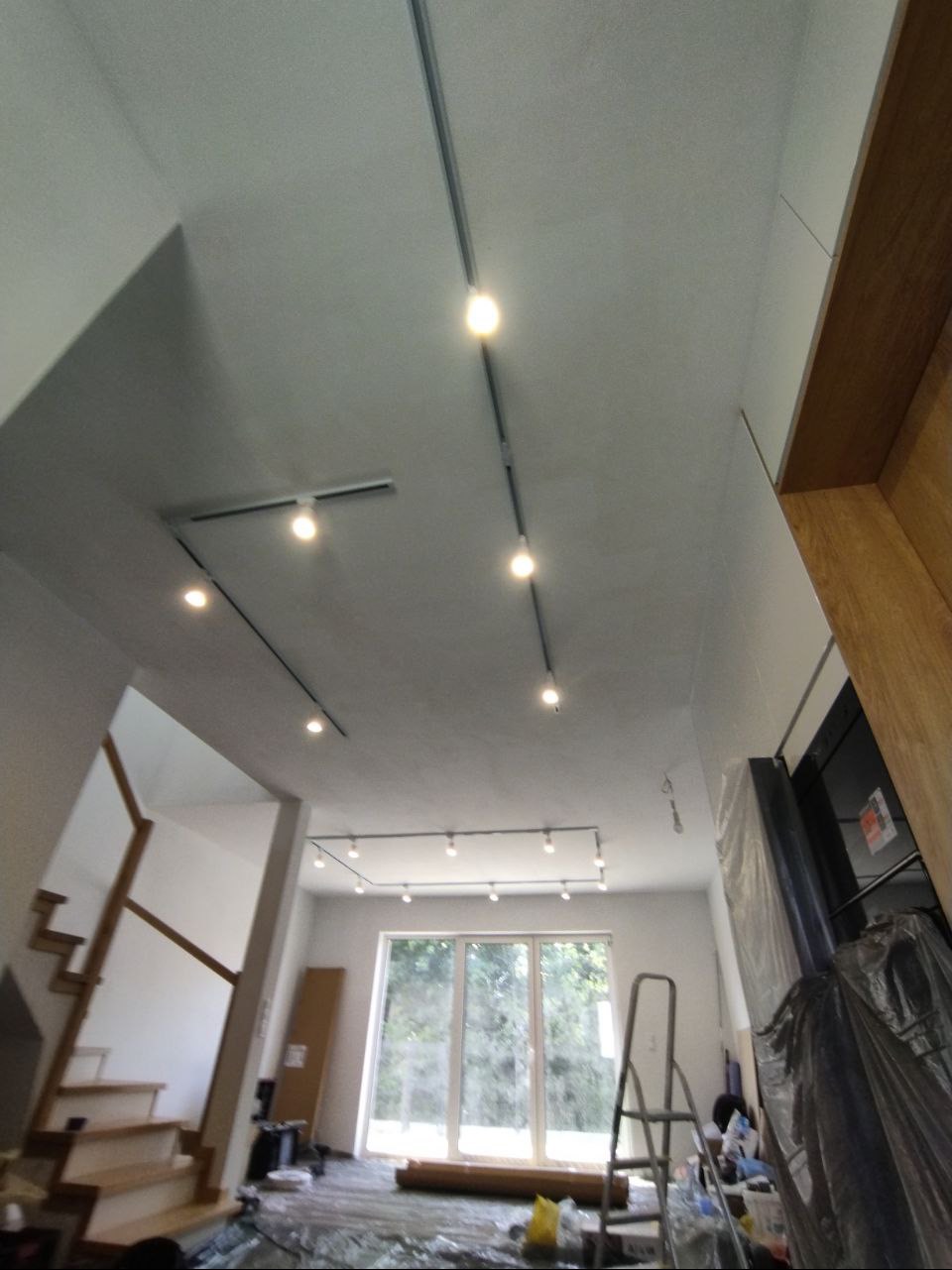 Szyny z zamontowanym oświetleniem punktowym na suficie w trakcie prac wykończeniowych; widoczne schody, okno i rozłożona folia ochronna.