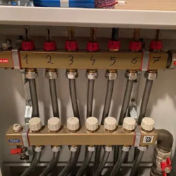 Rozdzielacz ogrzewania podłogowego z siedmioma obwodami, oznaczonymi cyframi od 1 do 7, z widocznymi przepływomierzami i zaworami regulacyjnymi, zamontowany w szafce instalacyjnej.