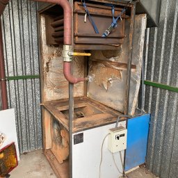 Zicher Technika Grzewcza Mateusz Dziembowski - Otwarty, skorodowany piec z widoczną instalacją, wentylatorem i przewodami, stojący przy blaszanym murze.