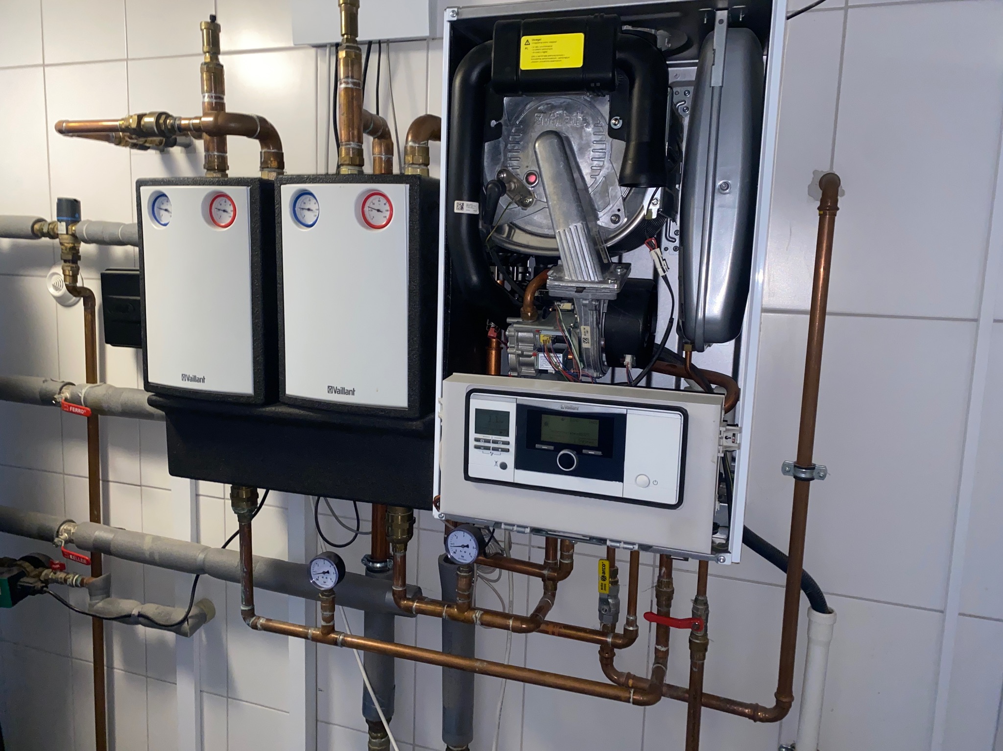 Otwarty piec gazowy marki Vaillant z widocznymi elementami wewnętrznymi i podłączonymi rurami miedzianymi oraz dwoma zewnętrznymi modułami z manometrami, zainstalowany na ścianie wyłożonej białymi...