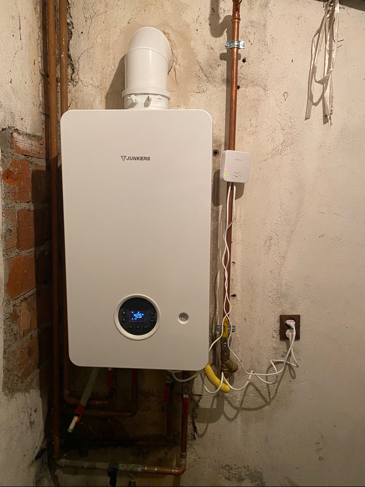 Biały, wiszący piec gazowy Junkers z wyświetlaczem 42.6 stopni Celsjusza, podłączony do instalacji miedzianej i gniazdka elektrycznego na tle surowej, betonowej ściany z fragmentem muru ceglanego.