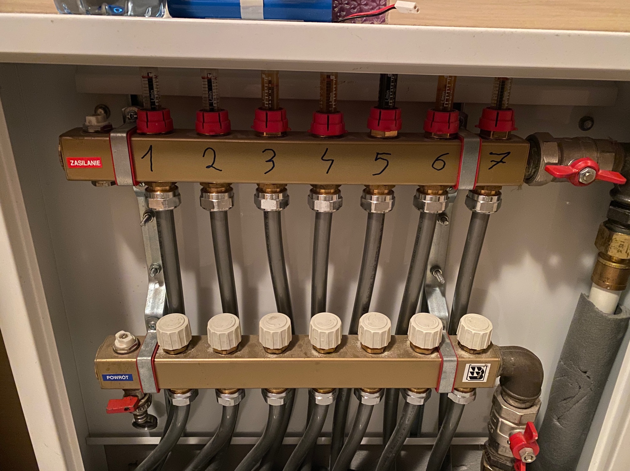 Rozdzielacz ogrzewania podłogowego z siedmioma obwodami, oznaczonymi cyframi od 1 do 7, z widocznymi przepływomierzami i zaworami regulacyjnymi, zamontowany w szafce instalacyjnej.