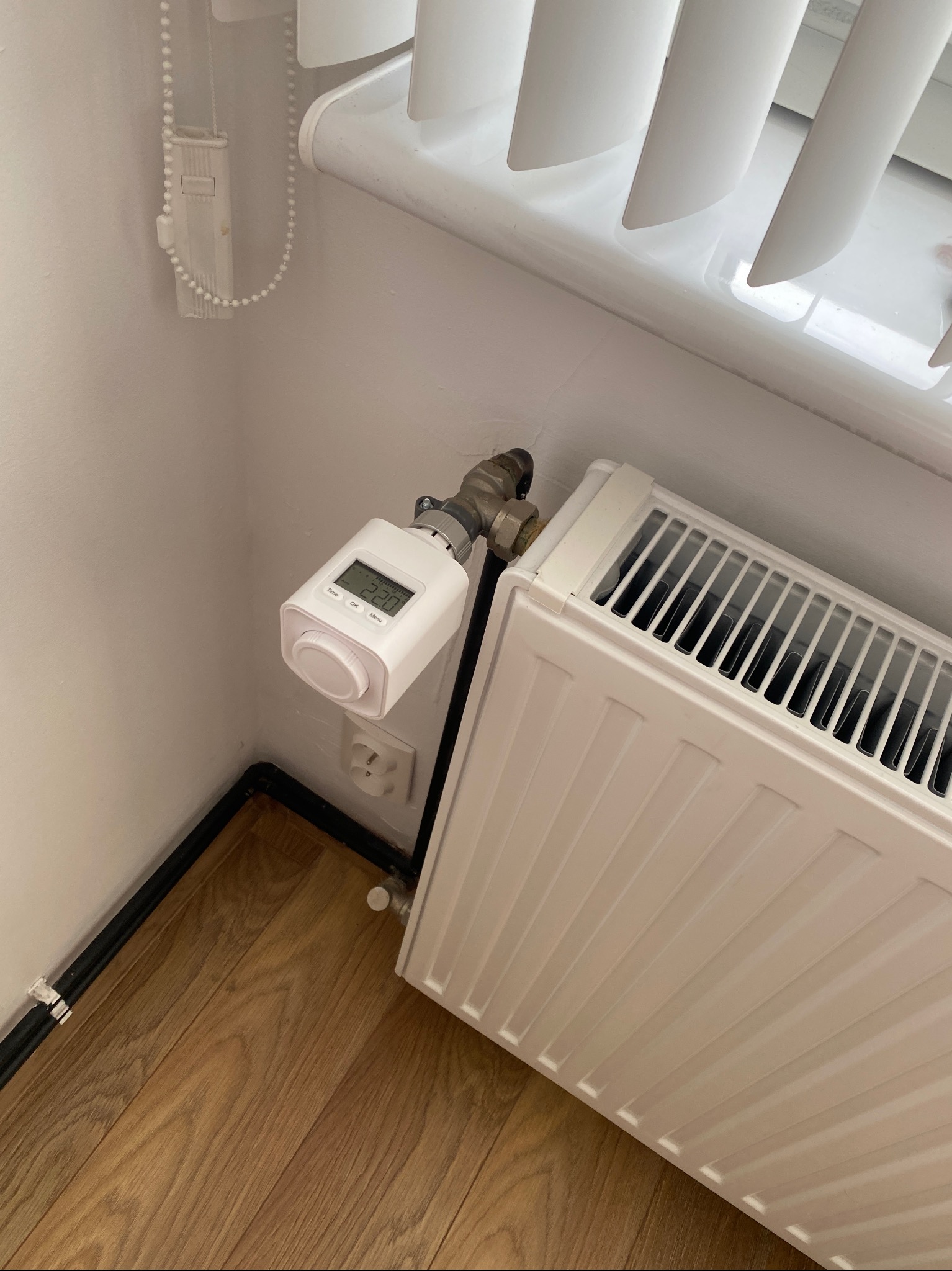 Biały grzejnik z elektronicznym termostatem pokazującym temperaturę 22 stopni Celsjusza, zamontowany przy oknie z żaluzjami i drewnianej podłodze.