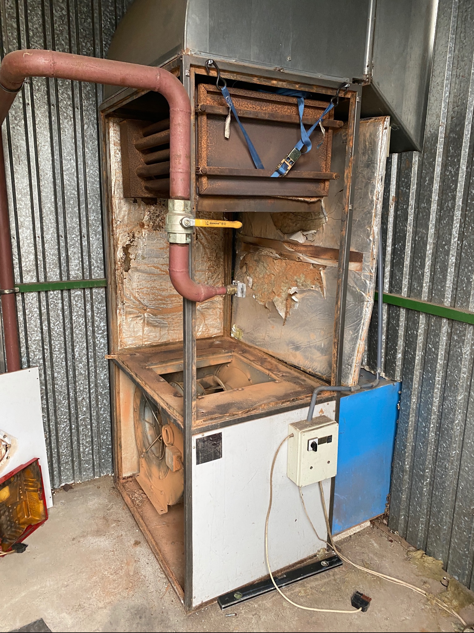 Otwarty, skorodowany piec z widoczną instalacją, wentylatorem i przewodami, stojący przy blaszanym murze.