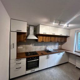 Paweł Nikiel - Nowoczesna kuchnia z białymi szafkami i drewnianymi akcentami, czarnymi uchwytami, płytą grzewczą i okapem. Jasne wnętrze z szarymi płytkami podłogowymi.