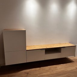 Paweł Nikiel - Nowoczesna, biała szafka RTV z drewnianym blatem, minimalistyczny design, oświetlenie punktowe, drewniana podłoga, jasne wnętrze.