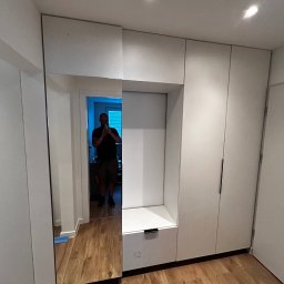 Paweł Nikiel - Biała szafa wnękowa z lustrem i siedziskiem w przedpokoju, widok z przodu, drewniana podłoga, oświetlenie sufitowe, odbicie osoby robiącej zdjęcie.