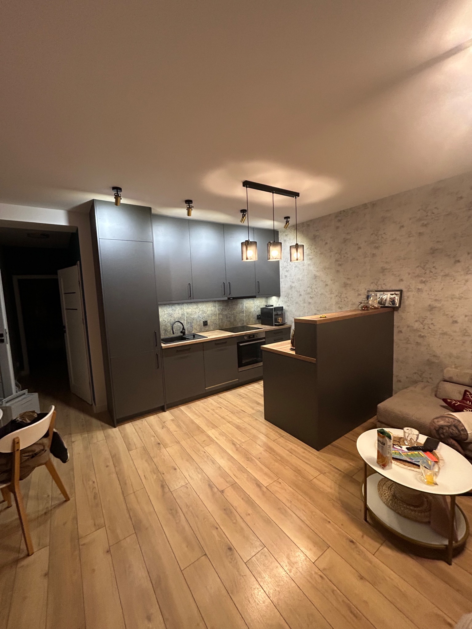 Nowoczesna kuchnia w odcieniach szarości z drewnianym blatem i podłogą. Widoczne oświetlenie punktowe i dekoracyjna ściana w tle. Minimalistyczny design.