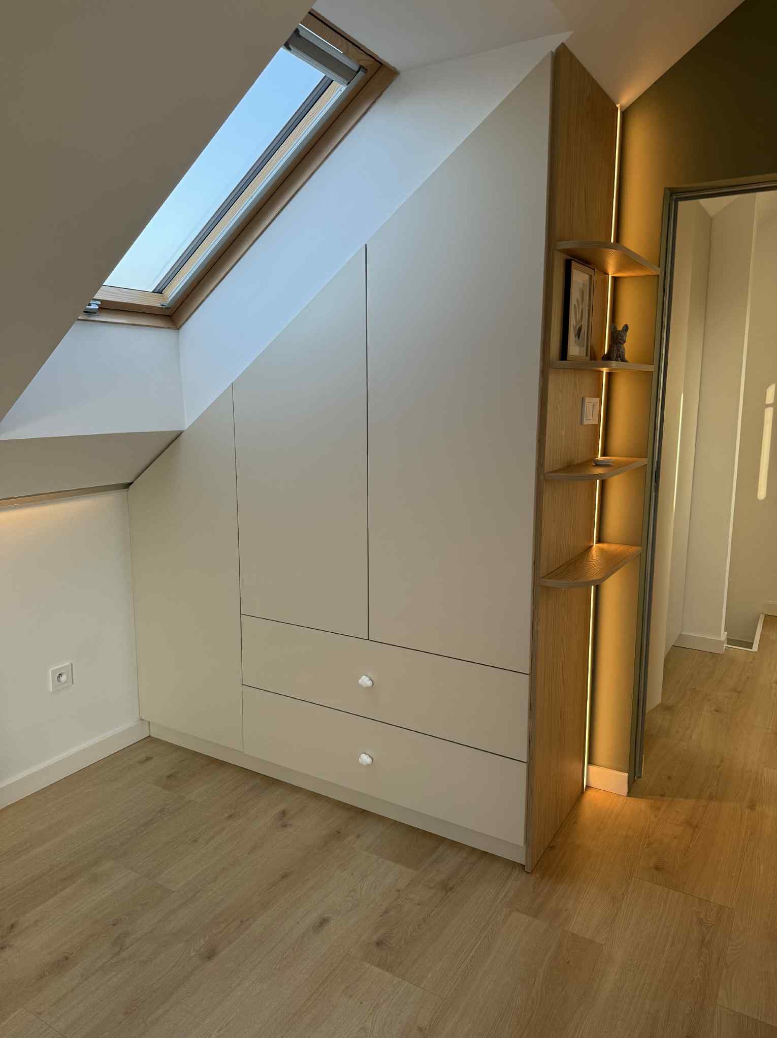 Biała szafa wnękowa z szufladami i drewnianymi półkami z oświetleniem LED, dopasowana do skosu dachu, na poddaszu z oknem dachowym i panelami podłogowymi.