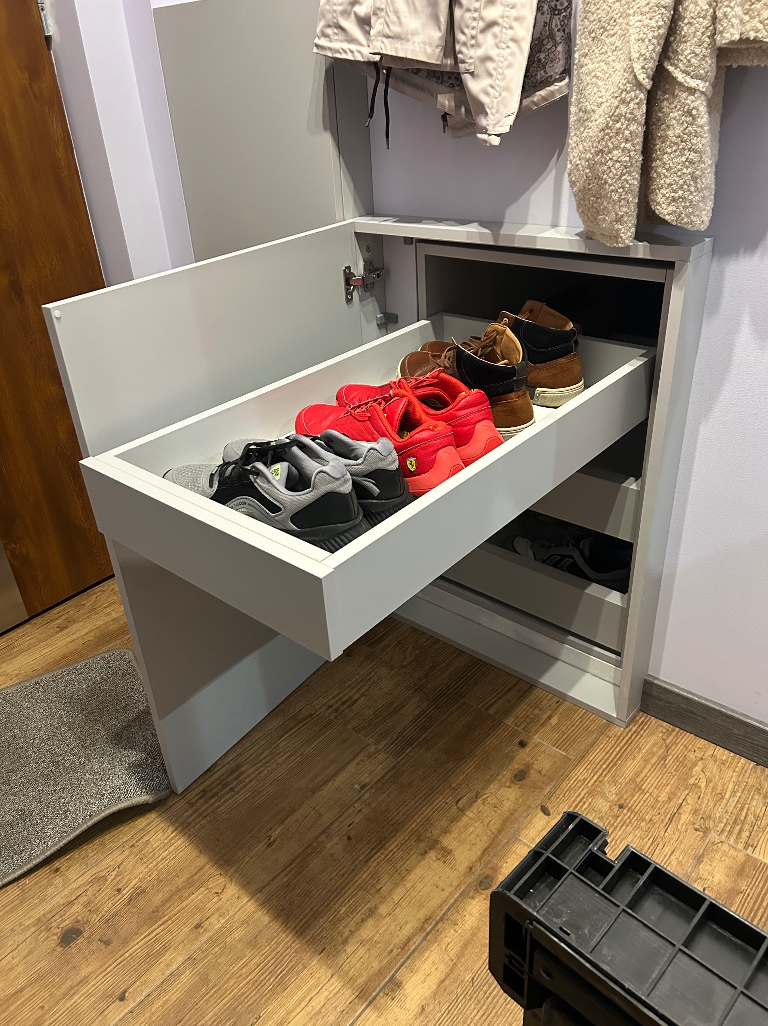 Nowoczesna, biała szafka na buty z wysuwaną szufladą, wypełniona różnorodnym obuwiem. Widoczne ubrania wiszące nad szafką i fragment drewnianej podłogi.