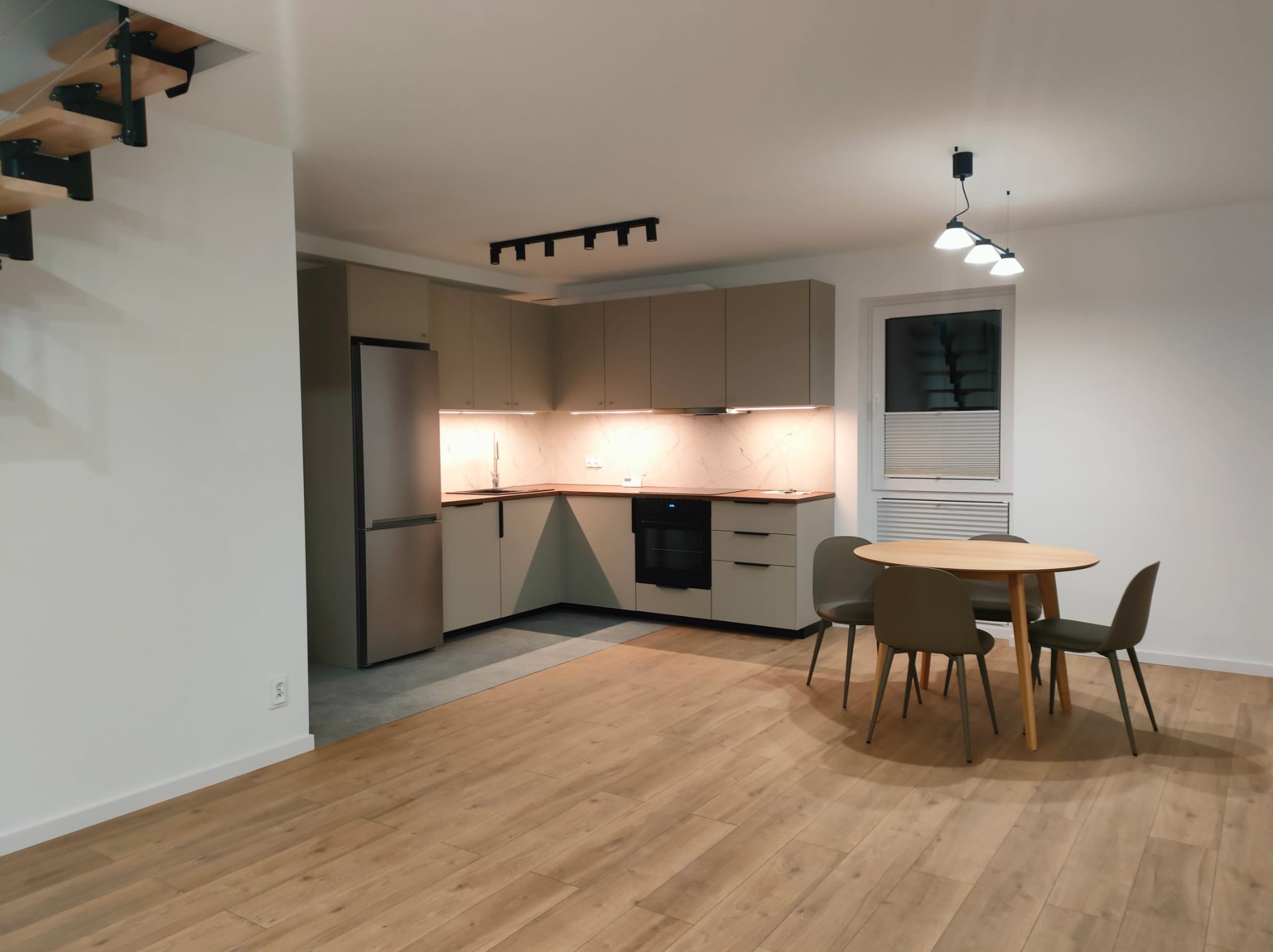 Jasna kuchnia z drewnianym parkietem i okrągłym stołem z krzesłami. Minimalistyczne oświetlenie sufitowe i schody z drewnianymi stopniami dodają wnętrzu nowoczesnego charakteru.