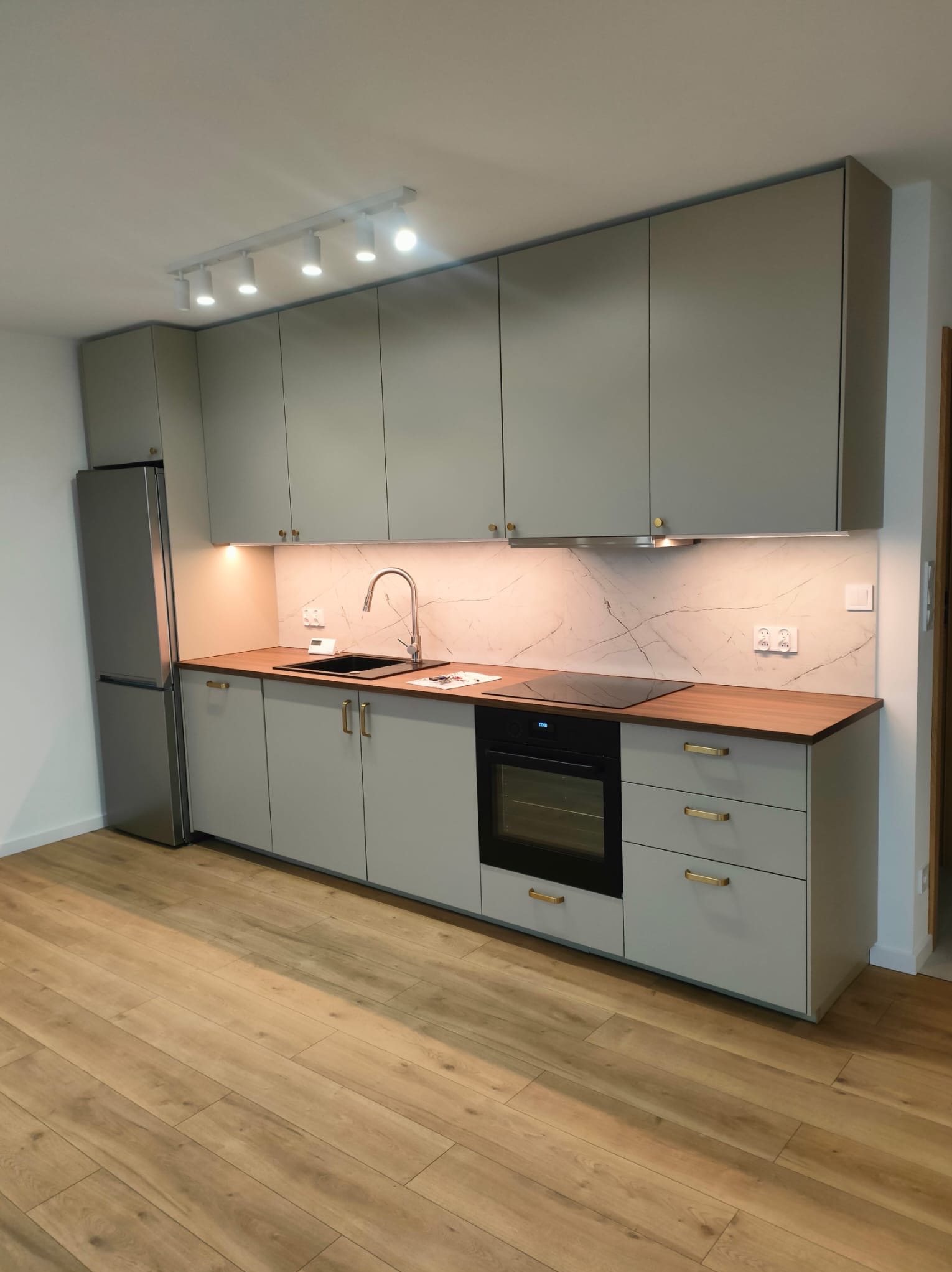 Nowoczesna kuchnia w odcieniach szarości z drewnianym blatem, oświetleniem punktowym i marmurowym backsplash, widziana z perspektywy podłogi z paneli.