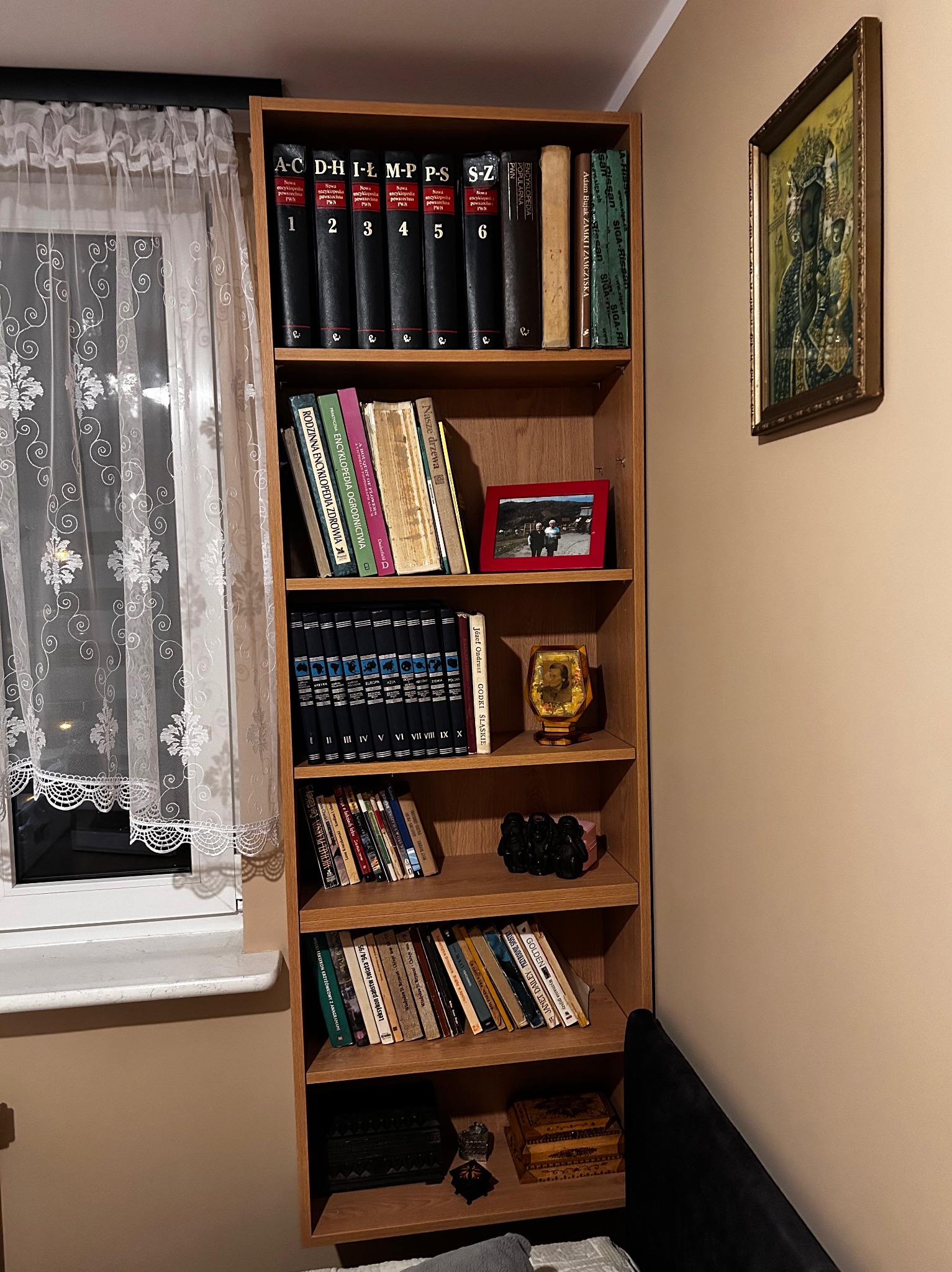 Drewniany regał z książkami, encyklopediami i pamiątkami, stojący przy oknie z koronkową firanką. Na ścianie wisi obraz religijny w złotej ramie.