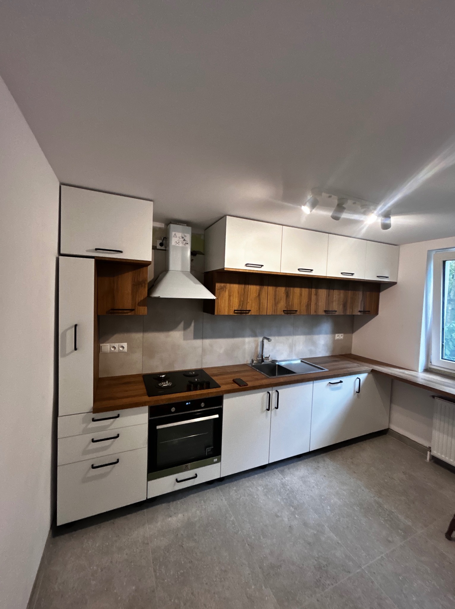 Nowoczesna kuchnia z białymi szafkami i drewnianymi akcentami, czarnymi uchwytami, płytą grzewczą i okapem. Jasne wnętrze z szarymi płytkami podłogowymi.