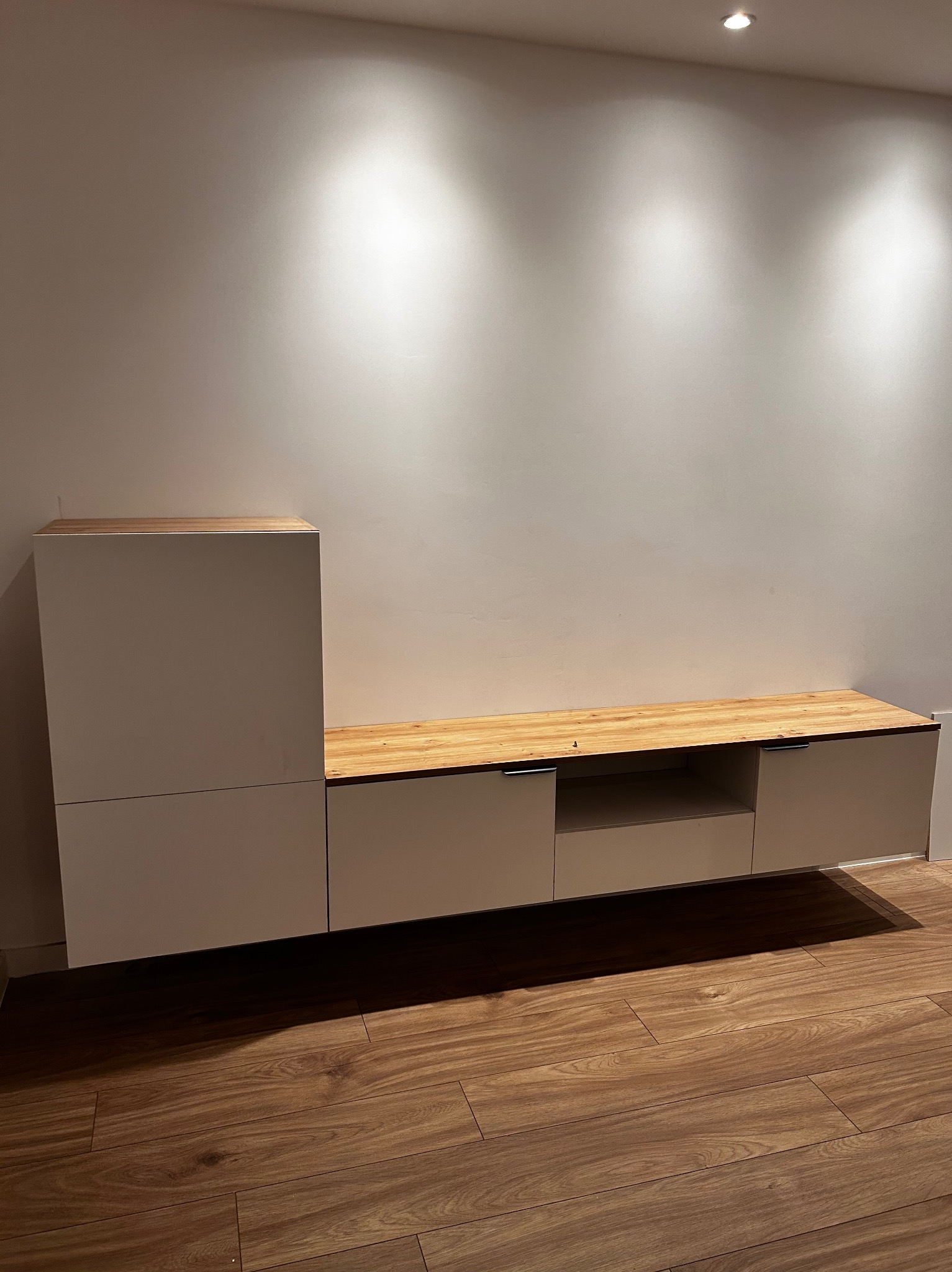 Nowoczesna, biała szafka RTV z drewnianym blatem, minimalistyczny design, oświetlenie punktowe, drewniana podłoga, jasne wnętrze.