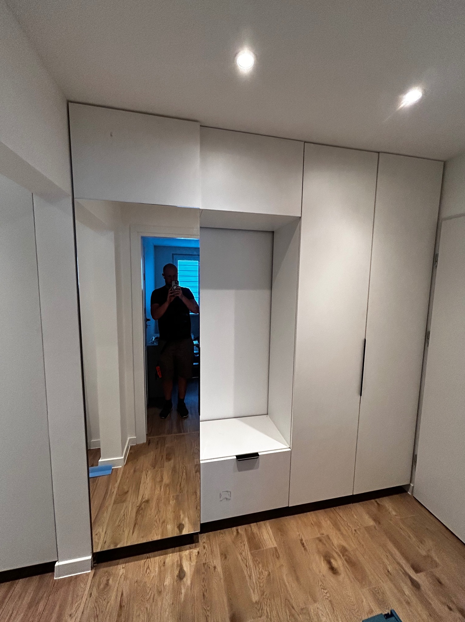 Biała szafa wnękowa z lustrem i siedziskiem w przedpokoju, widok z przodu, drewniana podłoga, oświetlenie sufitowe, odbicie osoby robiącej zdjęcie.