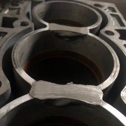 Mechanika Pojazdowa Serwis Chłodnic i Głowic Samochodowych - Fragment aluminiowego bloku silnika z widocznymi trzema cylindrami po naprawie, z wyraźnymi śladami spawania w miejscu uszkodzeń.