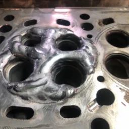 Głowica silnika samochodowego po spawaniu, widoczne ślady spawu wokół gniazd zaworowych, metalowa powierzchnia z otworami.