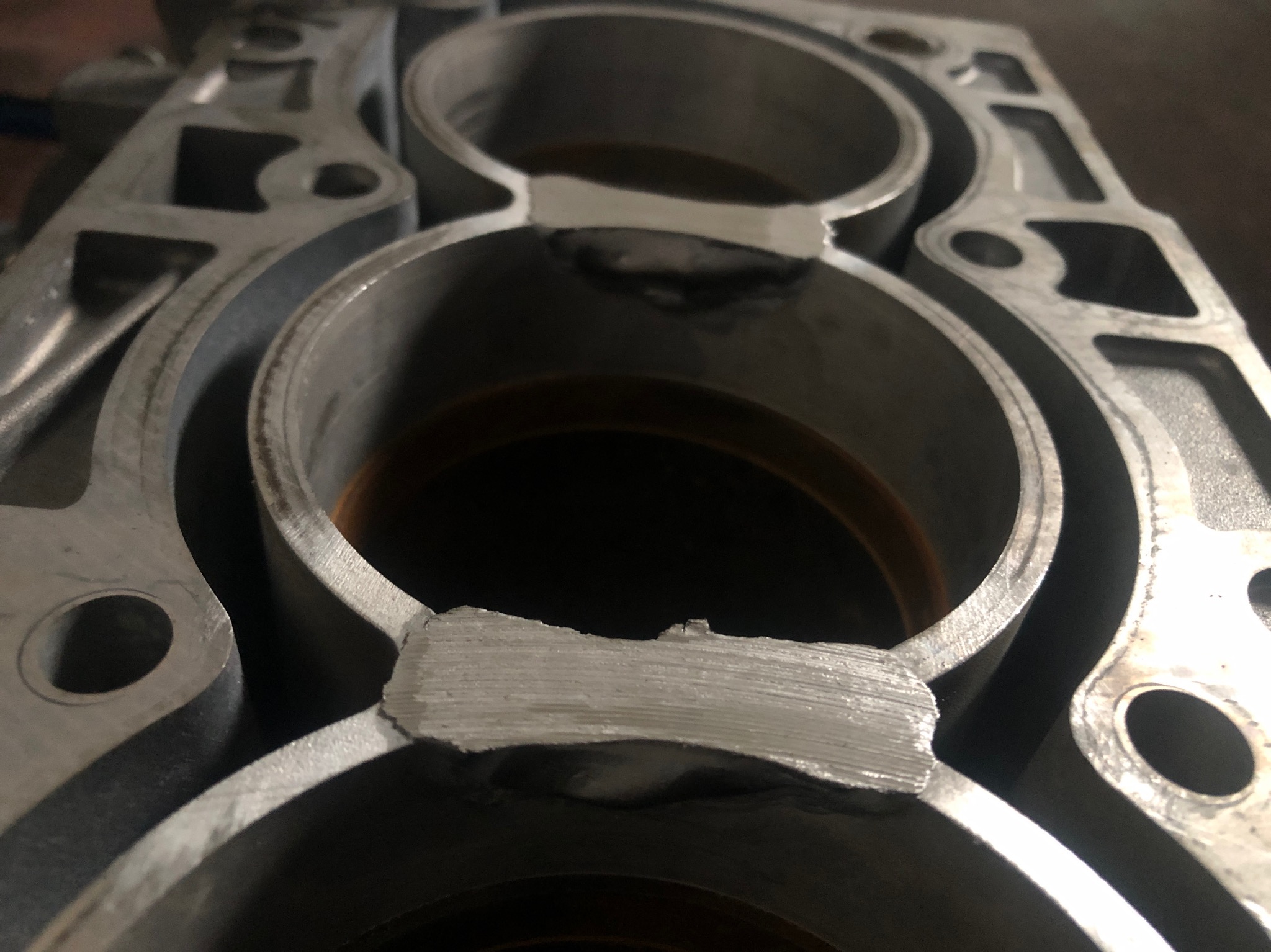 Fragment aluminiowego bloku silnika z widocznymi trzema cylindrami po naprawie, z wyraźnymi śladami spawania w miejscu uszkodzeń.