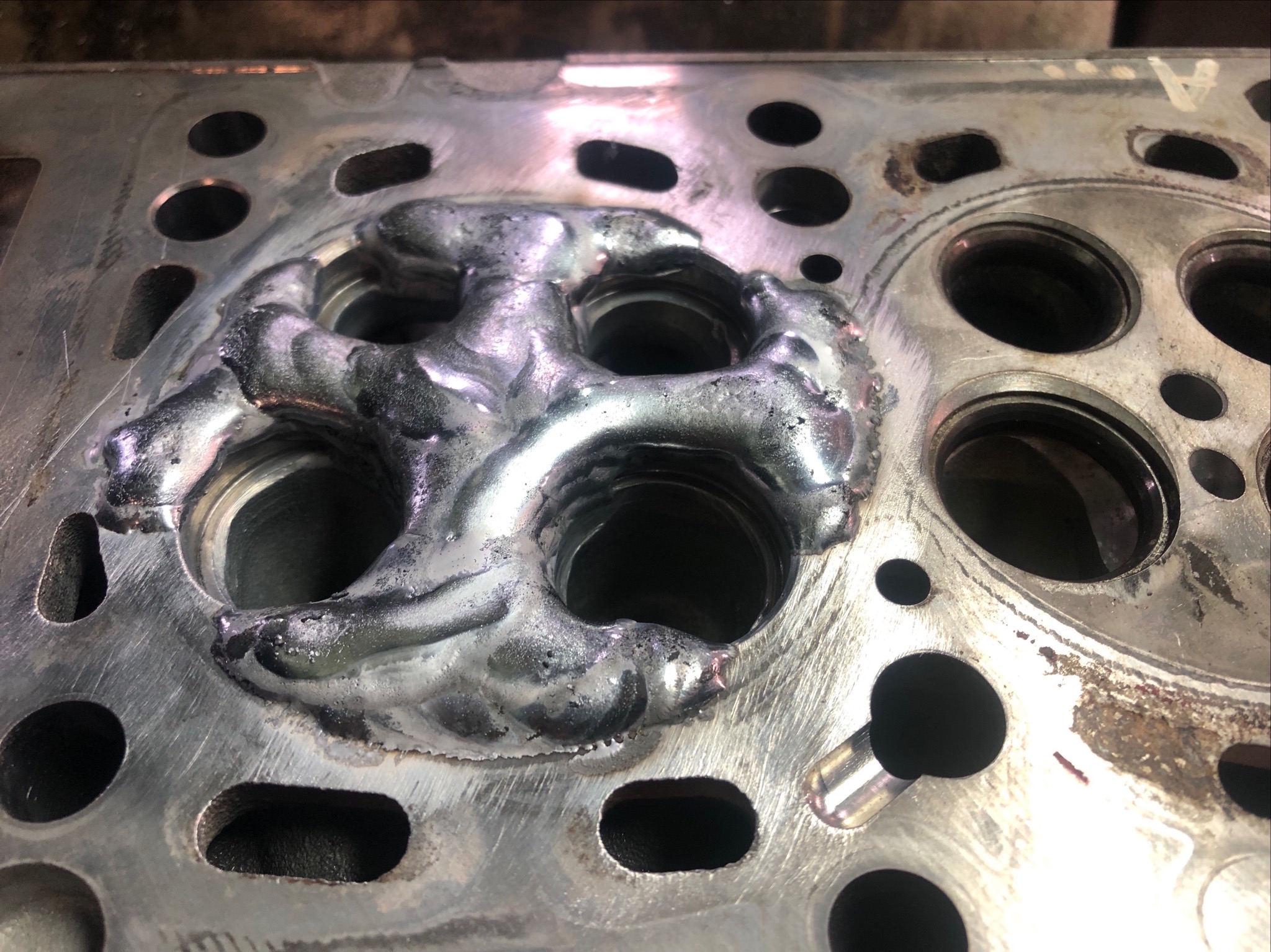 Głowica silnika samochodowego po spawaniu, widoczne ślady spawu wokół gniazd zaworowych, metalowa powierzchnia z otworami.