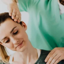 Kobieta poddawana terapii manualnej szyi przez terapeutę w zielonym uniformie; delikatny uśmiech pacjentki.
