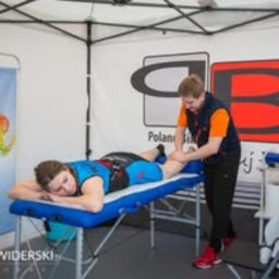 Przychodnia MOJA Ochota Warszawa - Fizjoterapia i Rehabilitacja - Mężczyzna w pomarańczowej koszulce i granatowej kamizelce wykonuje masaż nogi leżącej na niebieskim stole osobie w sportowym stroju, w tle biały namiot z logo.