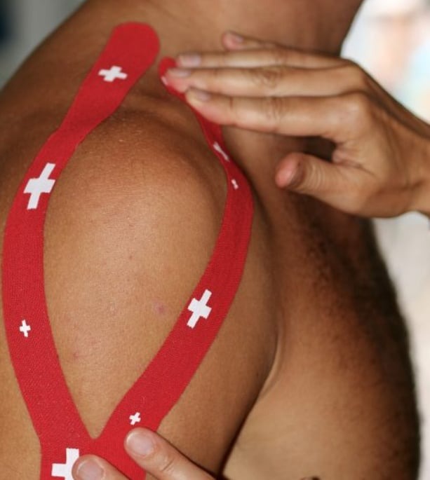 Aplikacja czerwonej taśmy kinesiotaping z białymi krzyżami na ramieniu i barku pacjenta, dotykana dłonią terapeuty.