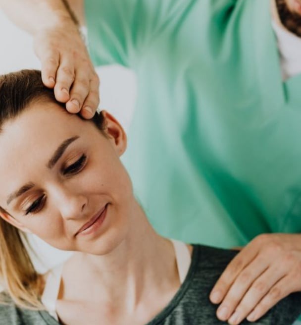 Kobieta poddawana terapii manualnej szyi przez terapeutę w zielonym uniformie; delikatny uśmiech pacjentki.