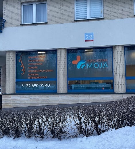 Fasada budynku przychodni rehabilitacyjnej z dużymi oknami, na których widnieją nazwy oferowanych usług: rehabilitacja, fizjoterapia, osteopatia, masaże, rehabilitacja dzieci, komora hiperbaryczna...