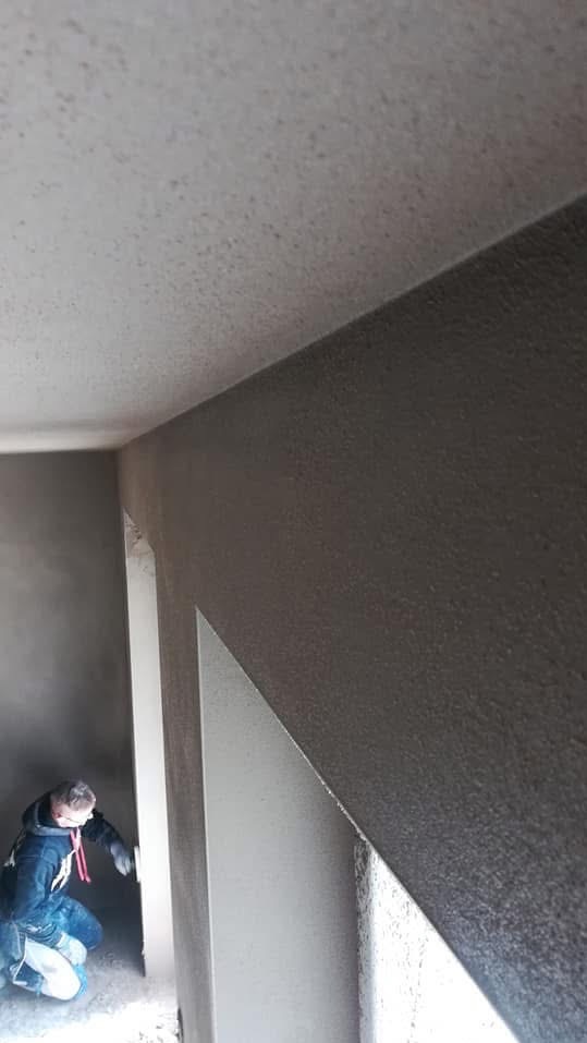 Tynkowanie ścian wewnątrz budynku. Widoczny pracownik w trakcie nakładania tynku na ścianę. Ściany w szarym kolorze, sufit biały. W tle widoczne kolejne pomieszczenie.