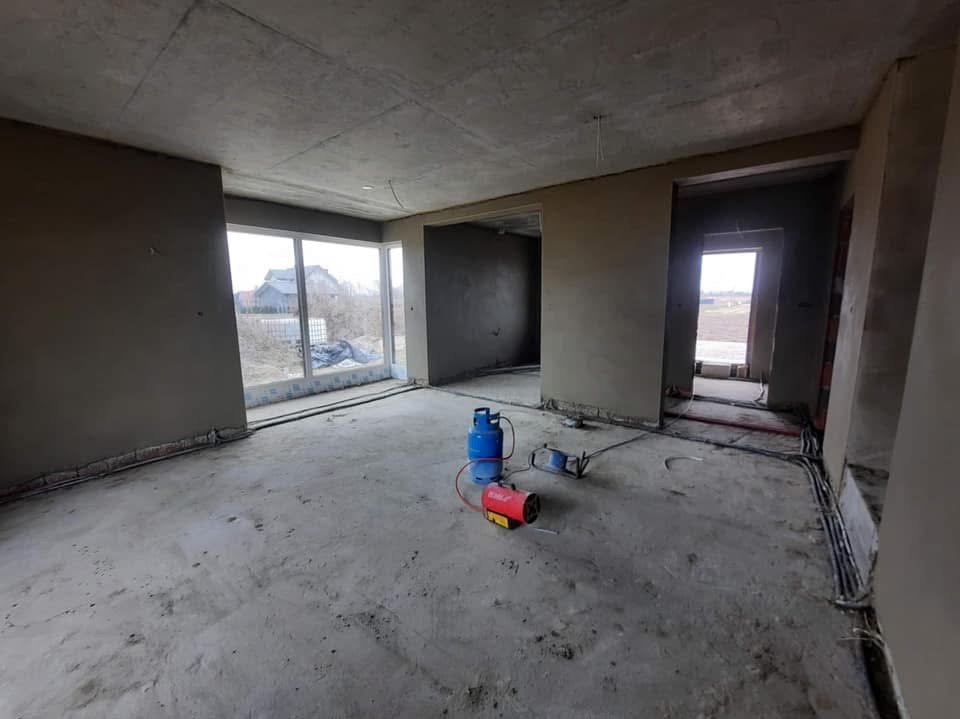 Surowe wnętrze w trakcie prac wykończeniowych: ściany pokryte świeżym tynkiem, widoczne instalacje elektryczne, butla gazowa i nagrzewnica. Duże okno z widokiem na zewnątrz.