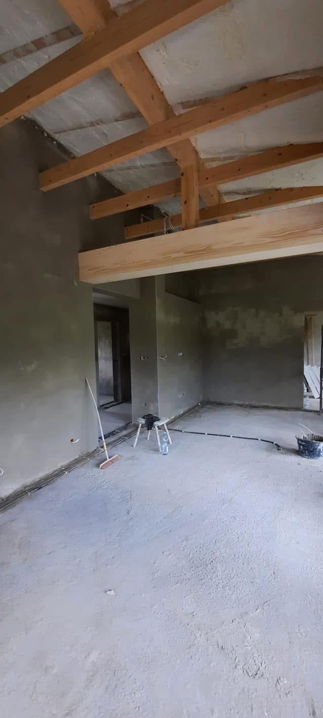 Surowe wnętrze po nałożeniu tynków, widoczne drewniane elementy konstrukcyjne dachu, ściany i podłoga pokryte świeżym tynkiem. W tle widoczne przejścia do innych pomieszczeń.