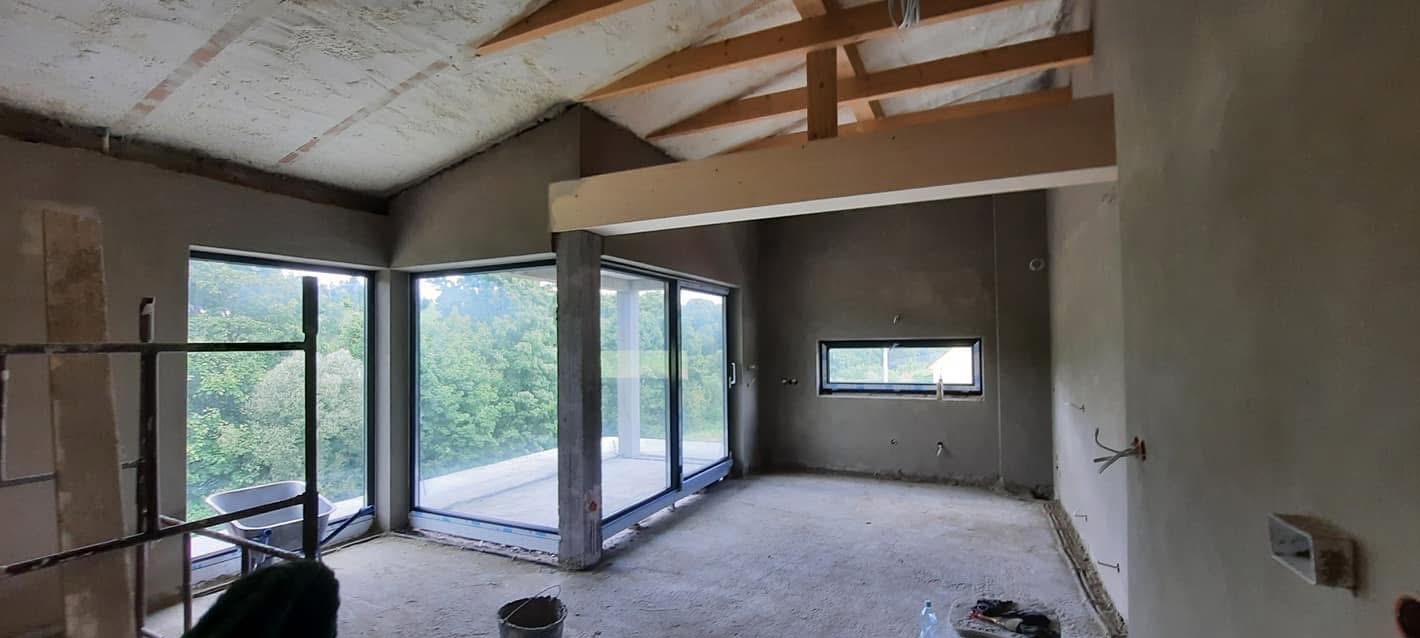 Wnętrze w trakcie prac wykończeniowych: surowe ściany, duże okna z widokiem na zieleń, drewniane elementy konstrukcyjne dachu. Widoczne ślady po pracach budowlanych.